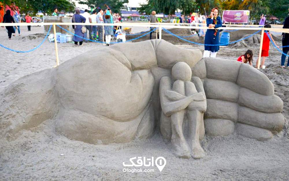 مسجمه شنی ساخته شده در کنار ساحل؛ یک دست شنی بزرگ پشت سر یک مجسمه شنی که پاهای خود را بغل کرده است