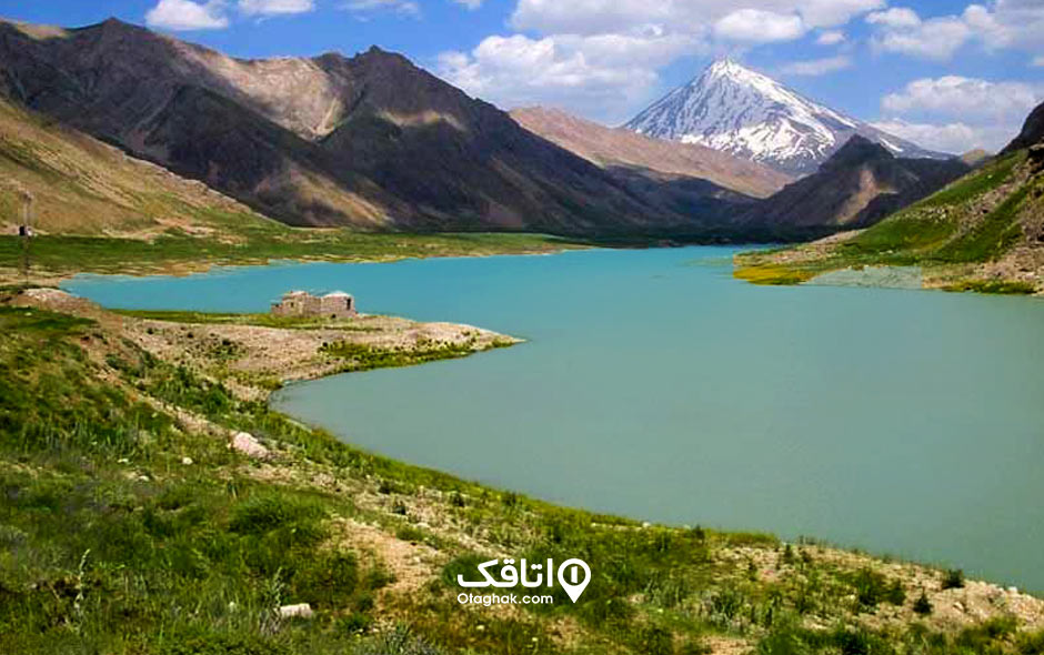 دریاچه سد به رنگ آبی که اطراف آن را گیاهان سبز پوشش داده‌اند و قله برف گرفته یک کوه نیز در دوردست‌ها پیداست