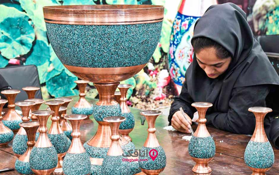 یک خانم در حال انجام فیروزه کوبی و تعدادی گلدان بزرگ و کوچک به رنگ مسی که فیروزه های آبی روی آن کوبیده شده است روی میز قرار دارند