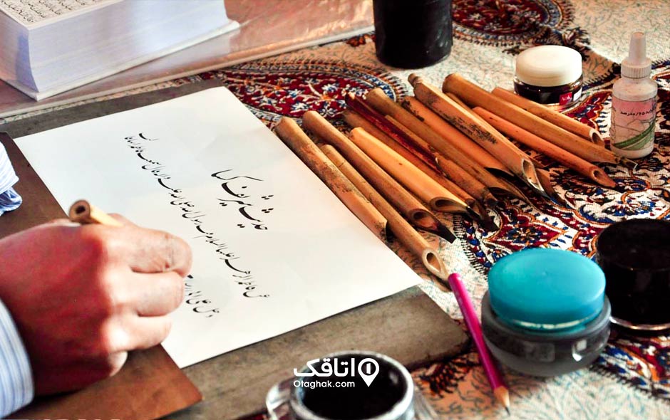 یک میز که روی ان تعدادی قلم خوش نویسی، مرکب و کاغذ است و دستان یک مرد در حال انجام خوش نویسی