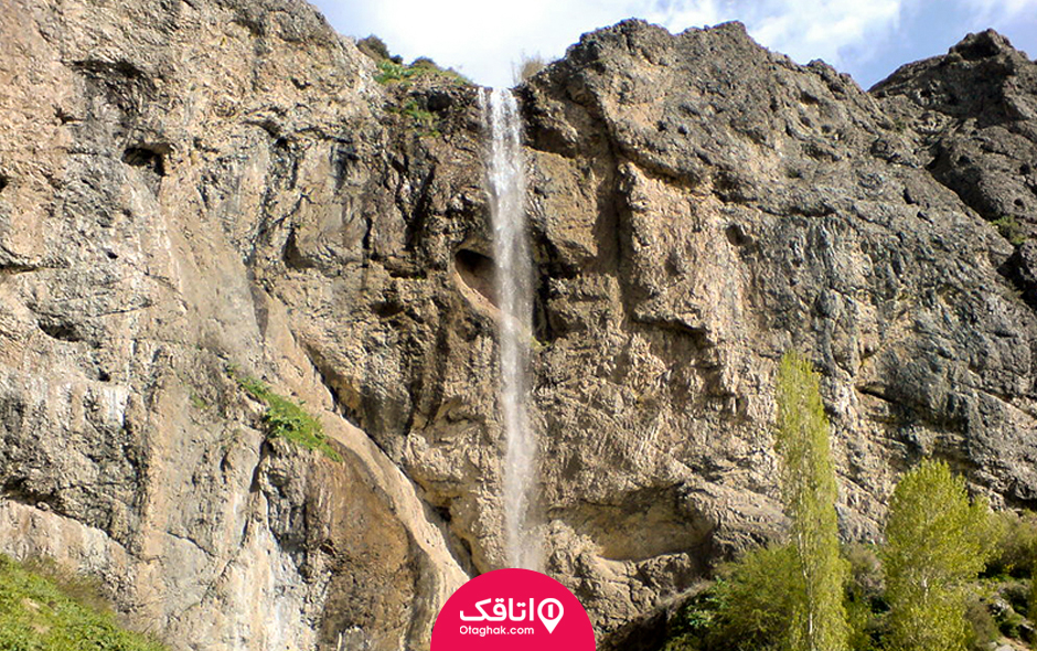 آب در حال جاری شدن از کوهی بلند و درختان بلندی در پایین آبشار که قسمتی از سر آن ها پیداست