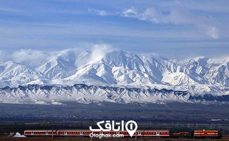 کوهستان پر از برف که روبروی آن یک ریل راه آهن قرار دارد و قطار قرمز رنگ در حال عبور است