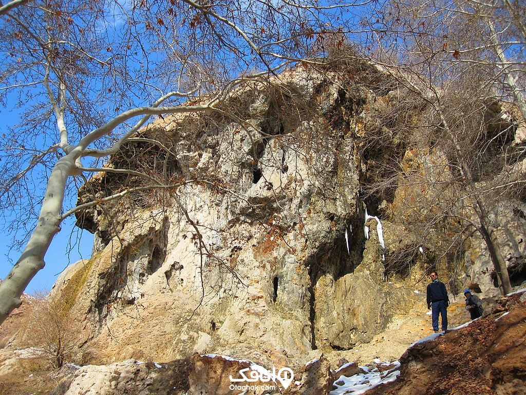 یک کوه و آبشار یخ زده در میان درختان بدون برگ و یک نفر که جلوی آن ایستاده