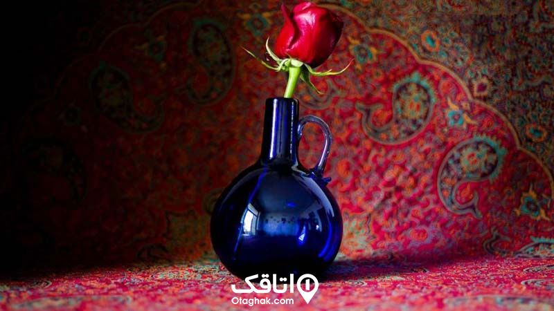 یک گلدان شیشه ای آبی رنگ که یک گل شاخه گل قرمز درون آن قرار دارد روی یک ترمه 