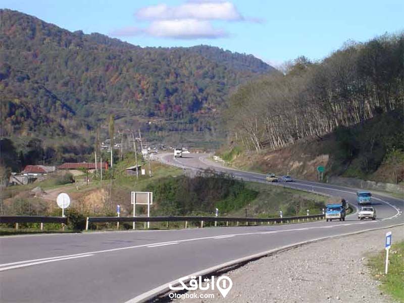 جاده ای دو طرفه و کوهستانی که کوه و درختانی در کنار ان دیده می شود و ماشین هایی در حالغبور از جاده