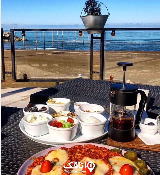یک میز صبحانه آماده شده با چای، املت، گوجه و خیار، خرما، پنیر و... نزذیک به ساحل با نمای دریا
