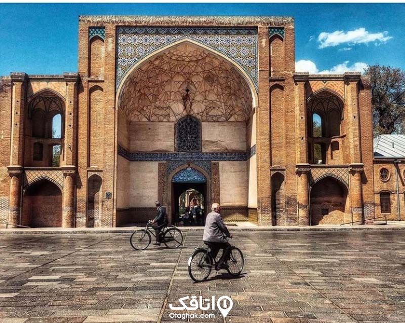 دو مرد با دوچرخه در حال گذشتن از جلو ورودی شبستان مانند مسجدی با گچ کاری و کاشی کاری