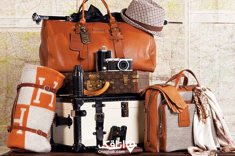 یک عدد چمدان قدیمی سفید با دسته و کمربندهای سیاه، یک چمدان کوچک قهوه ای روی آن قرار دارد، بالای آنها یک ساک قهوه ای، یک کلاه و یک دوربین قدیمی قرار گرفته در کنار این وسایل یک کیف دستی زنانه طوسی-قهوه ای و یک پت. مسافرتی قوه ای سفید قرار دارد