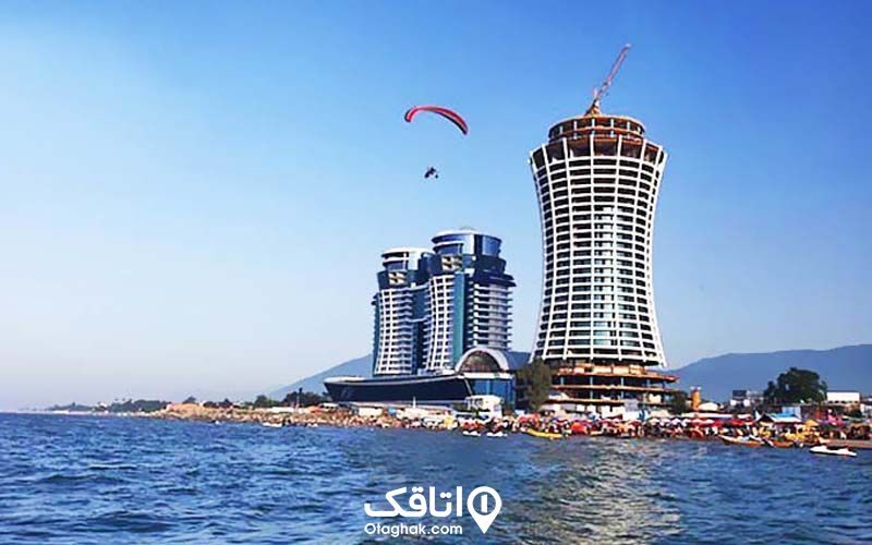 ساحل کنار برج های تجاری متل قو، قایق های تفریحی لب ساحل و یکنفر در حال چتر بازی روی دریا
