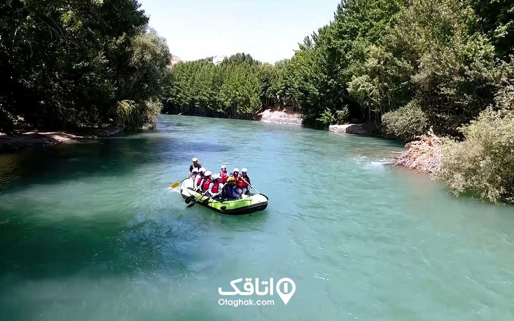 نه نفر داخل یک قایق رفتینگ در میان رودخانه ای آب خروشان احاطه شده با درختان انبوه سبز رنگ