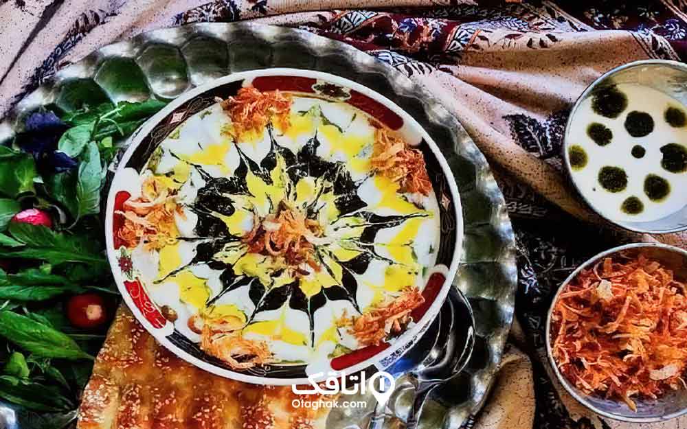 یک کاسه آش داخل سینی به همراه نان بربری و سبزی خوردن و یک ظرف سیر سرخ شده 