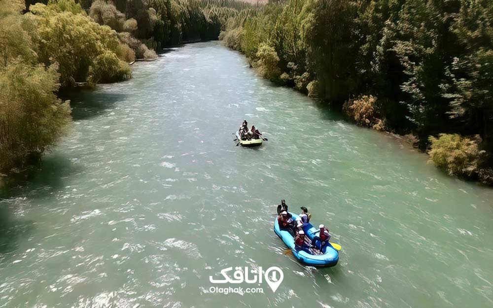 چندین نفر داخل دو قایق رفتینگ میان روخانه ای که دو طرف آن درختان سرسبز است.