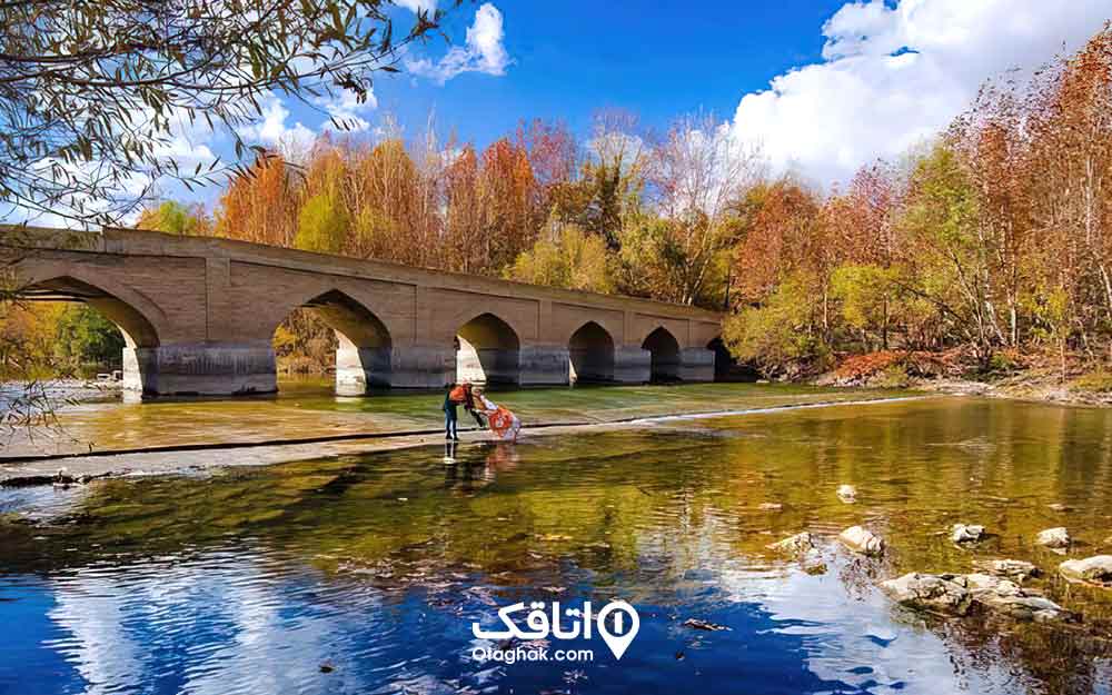 یک پل بر روی رودخانه ای آرام با درختان رنگارنگ پاییزی و آسمان ابی