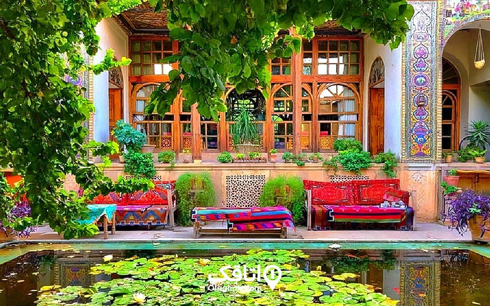 خانه اب معماری سنتی و در و پنجره هایی چوبی و حوض و درختی بزرگ و سبز رنگ در حیاط