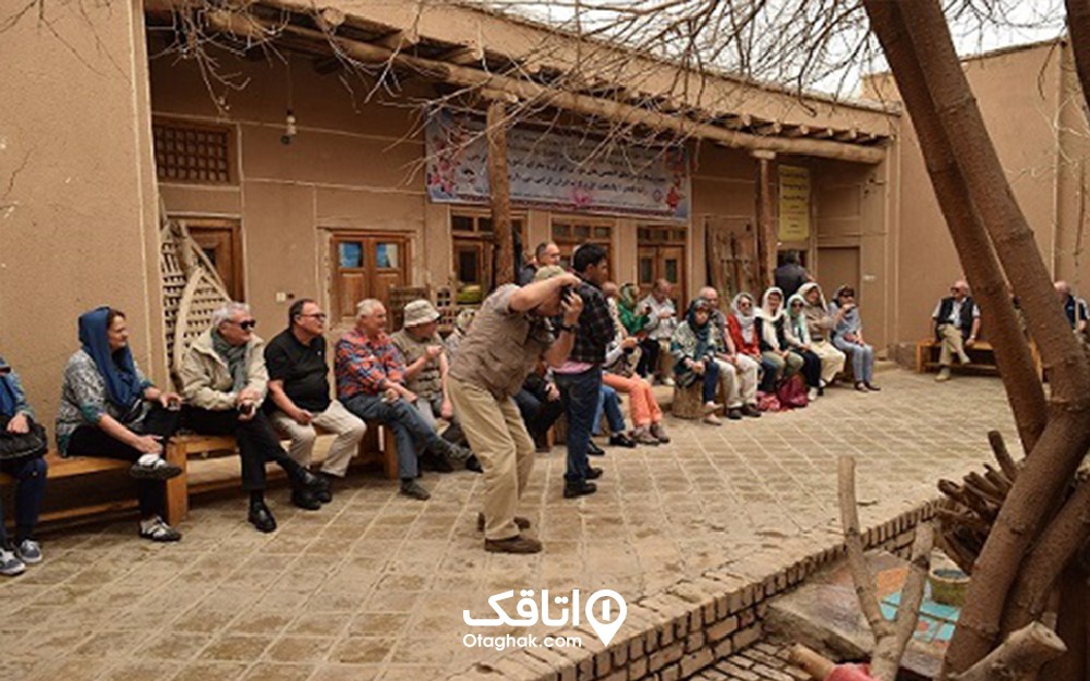 ساختمانی گلی و مردمی که جلوی آن نشسته اند و تعدادی از افراد که ایستاده اند