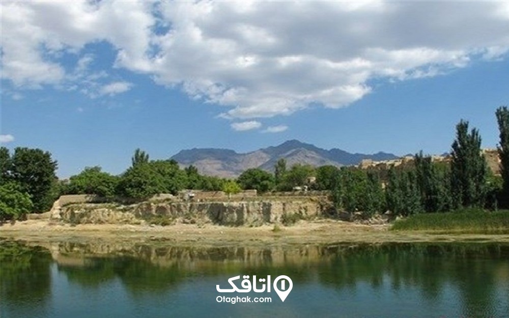 دریاچه ای آرام که در کنار ان درختان دیده می شوند و کوه هایی در دور دست زیر آسمان آبی